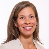 A woman with long brown hair, wearing a light-colored blazer and hoop earrings, smiling at the camera against a plain white background.