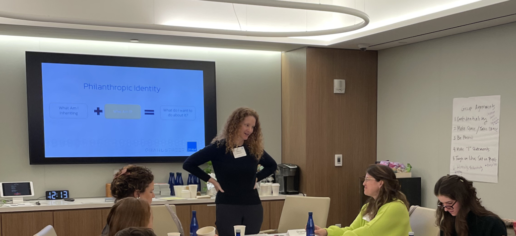 A woman stands and speaks to three seated women around a table in a modern conference room. A presentation slide titled Philanthropic Identity is displayed on a large screen behind her.